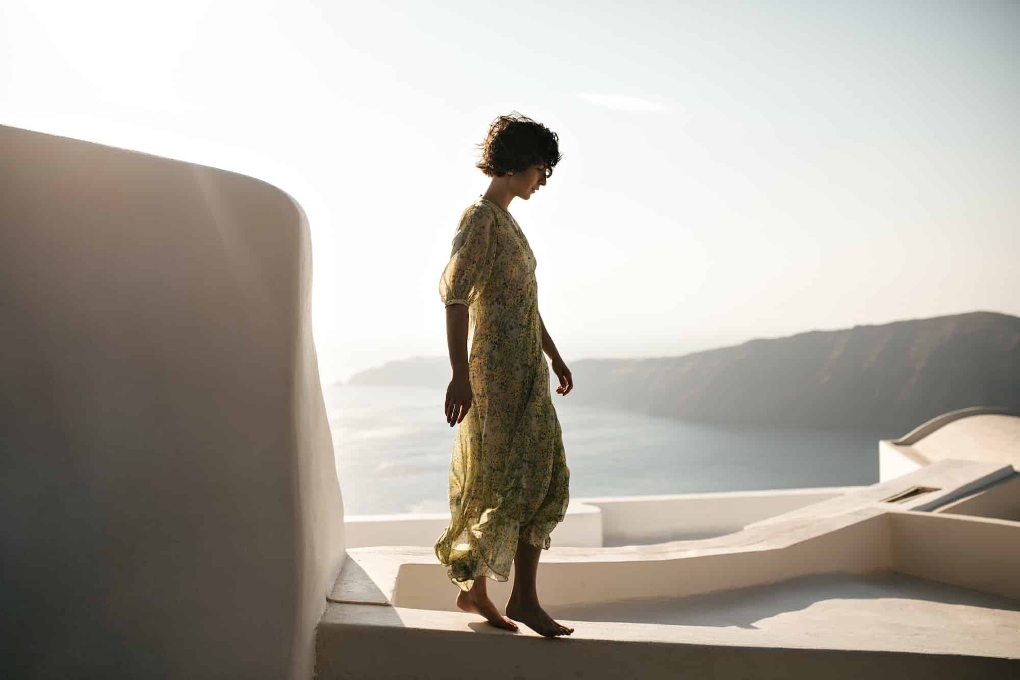 Full-length portrait of brunette woman in midi dress walks on terrace with sea view. Curly short-ha