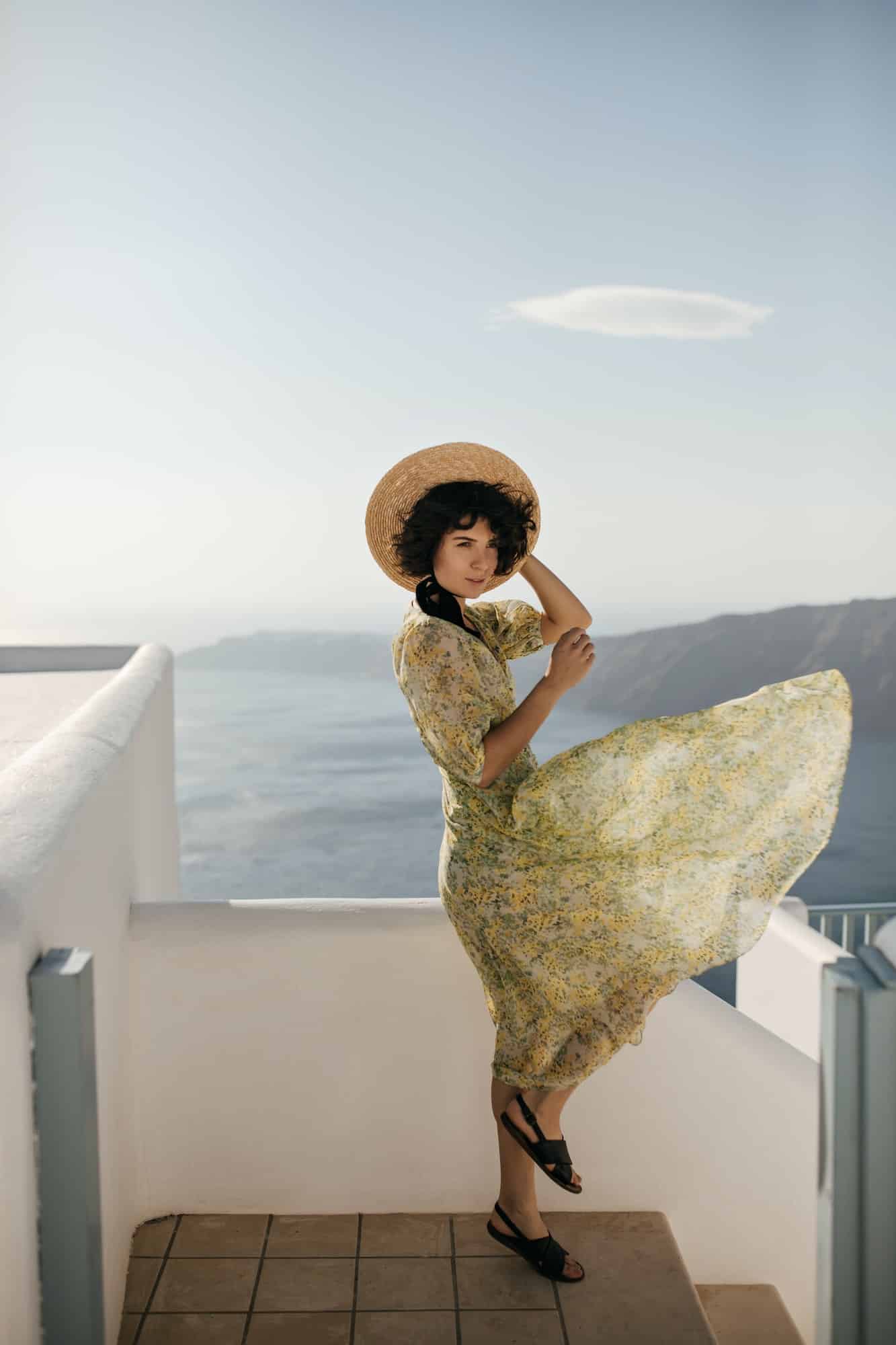 Attractive woman in yellow midi dress coquettishly poses on sea background. Brunette lady in hat an
