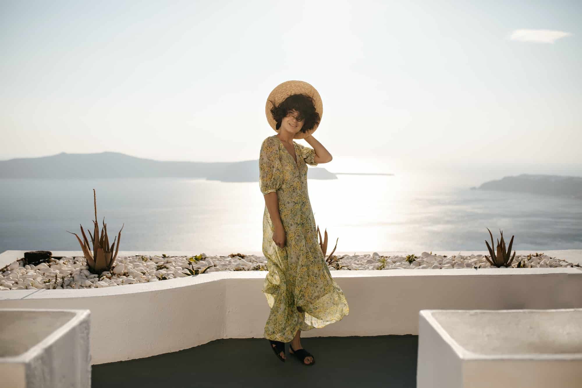 Full-length portrait of happy brunette lady in midi dress and boater posing on terrace with sea vie