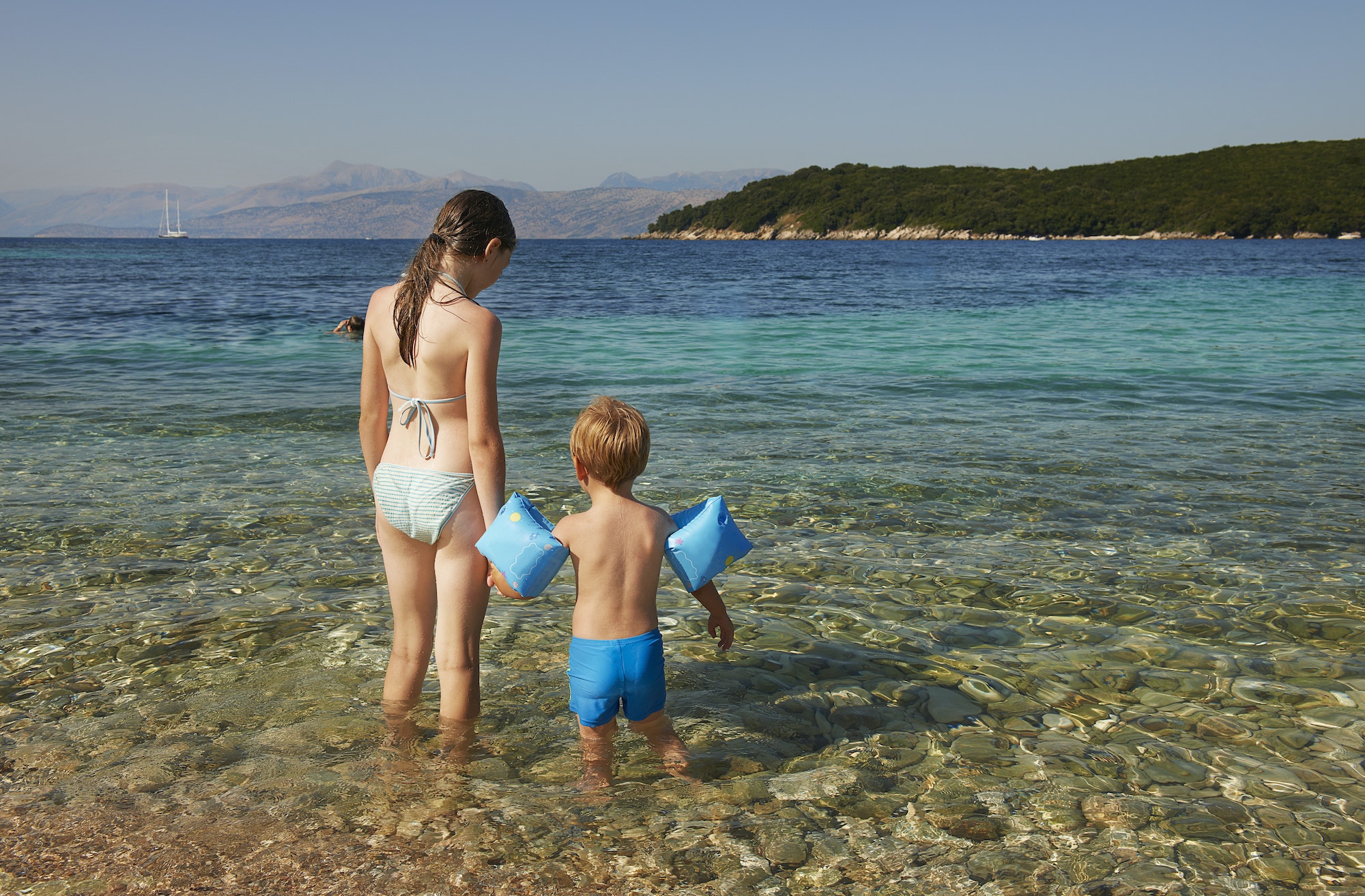 Rear view of girl and boy wearing blue armbands paddling in sea, Corfu, Greece