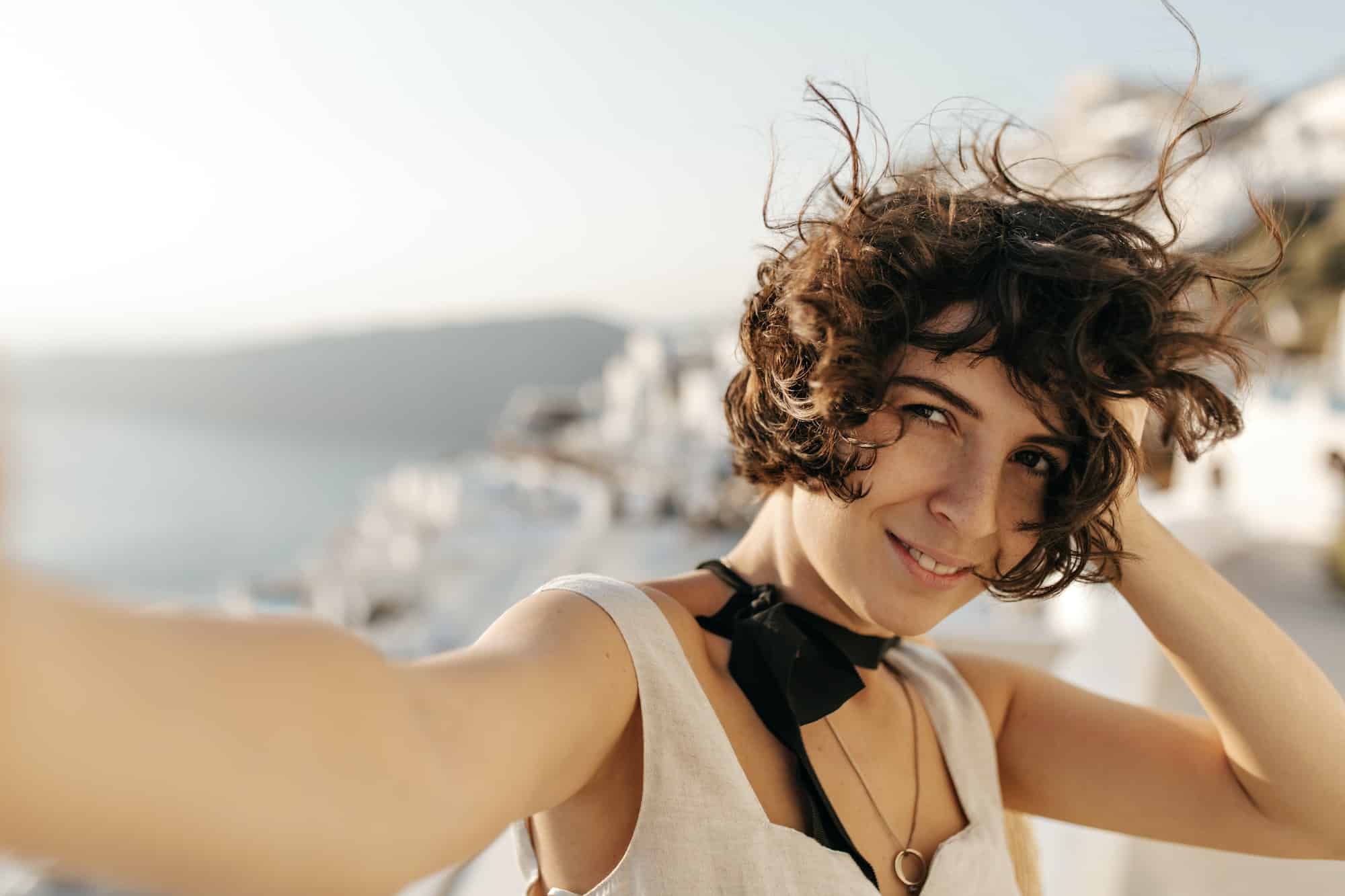 Brown-eyed curly woman in good mood takes selfie in beautiful place with sea view. Brunette attract