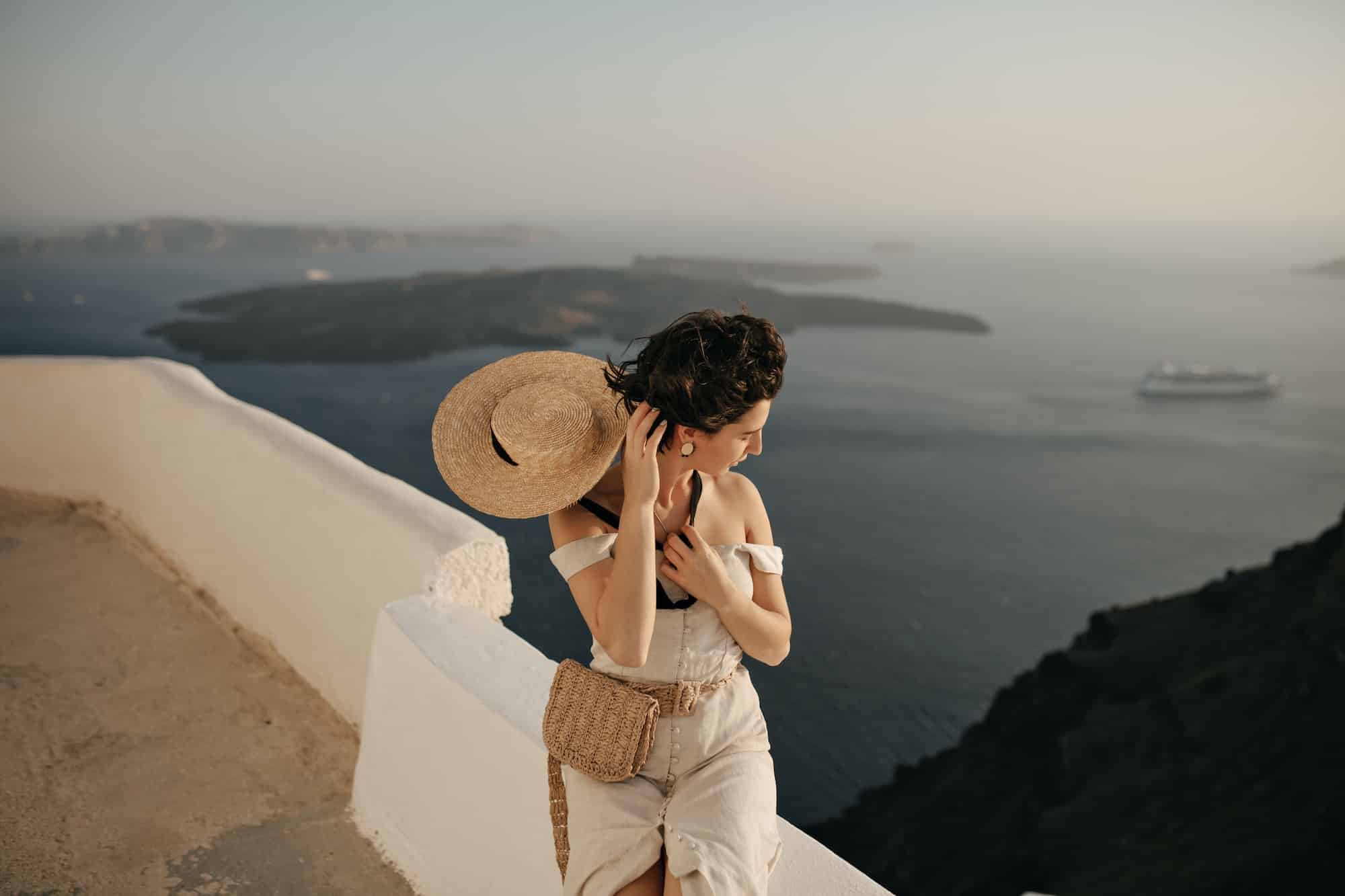 Young tanned lady in elegant beige dress holds straw boater. Brunette woman sits on white fence and