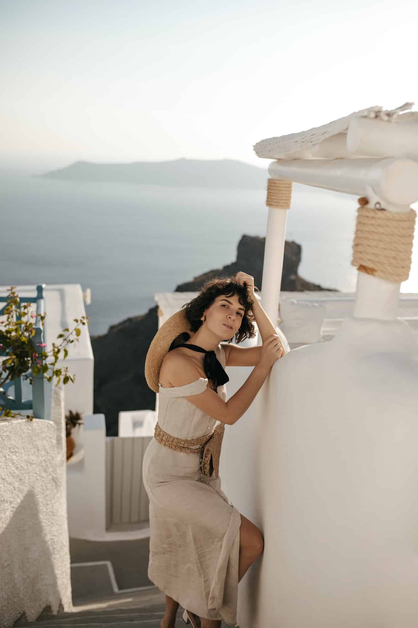 Pretty lady in beige midi dress and with straw bag leans in white wall and looks into camera outsid