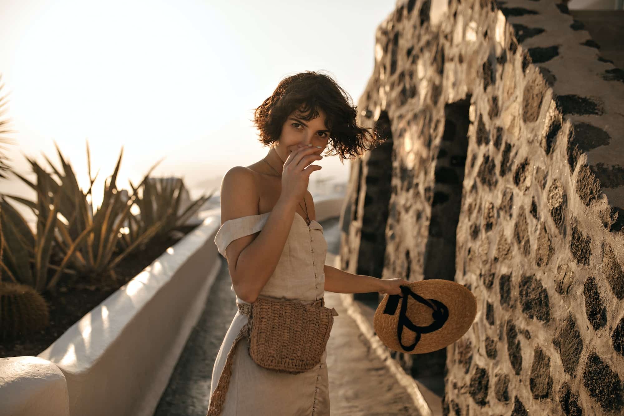 Brown-eyed brunette woman in beige dress with straw bag holds wide-brimmed hat and moves along path