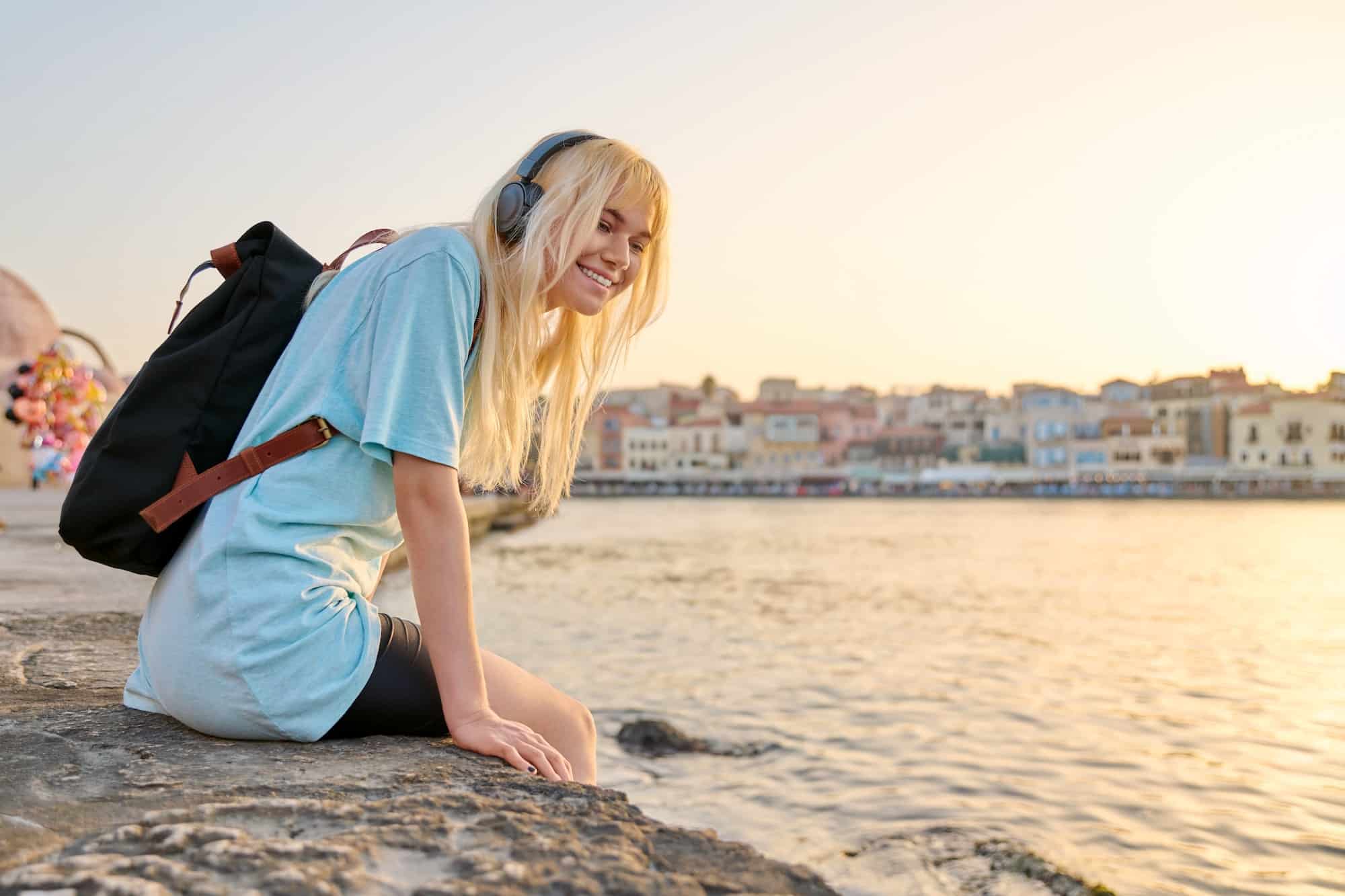 Happy teenage female in headphones with backpack enjoying tourist European city, seascape